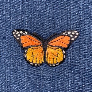 May include: Orange and black embroidered butterfly patch with white accents. The patch is on a blue denim background.