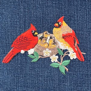 May include: Embroidered patch featuring two red cardinal birds near a nest with baby birds. The patch includes a branch with green leaves and white flowers. The patch is on a blue denim background.