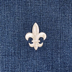 May include: A cream-colored fleur-de-lis patch is centered on a blue denim background. The patch is embroidered with a detailed design, showcasing the classic three-petal shape. The denim has a textured appearance.