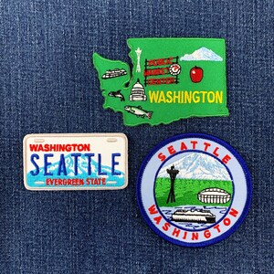 May include: Three embroidered patches featuring Washington state themes. One is a green map of Washington with landmarks and the text "PUBLIC MARKET CENTER" and "WASHINGTON". One is a rectangular license plate patch reading "WASHINGTON SEATTLE EVERGREEN STATE". One is a circular patch with "SEATTLE WASHINGTON" text.