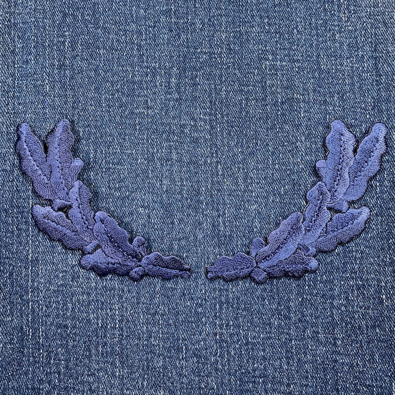 Pu&ograve; includere: Due applicazioni in tessuto ricamato blu a forma di corona d'alloro. Le applicazioni sono su uno sfondo di denim blu.