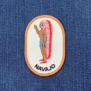 May include: A brown oval embroidered patch with a white background. The patch features a Native American man wearing a red and white feathered headdress. The text "NAVAJO" is embroidered in black below the image.