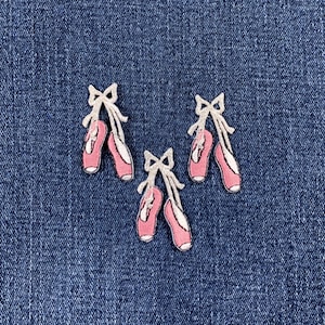 May include: Three embroidered patches of pink ballet shoes with white bows and ribbons. The shoes have a white outline, set against a blue denim background.