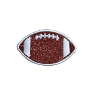May include: An oval-shaped football patch featuring a dark brown glittery texture. The design includes white embroidered stitching details and white vertical stripes on both ends, all outlined with a white stitched border for a clean finish.