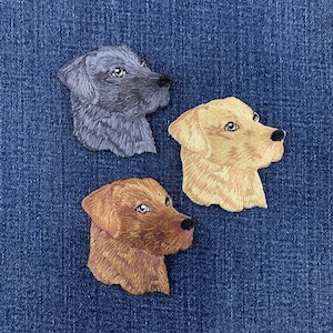 May include: Three embroidered dog head patches featuring detailed stitching. One patch is black, one is golden yellow, and one is a reddish-brown color. Each dog is depicted in profile facing right, showcasing realistic fur texture and dark noses.