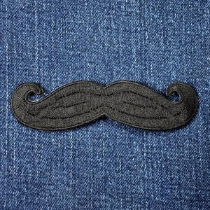 May include: A black embroidered mustache patch with a curved design, set against a blue denim background. The patch features detailed stitching and a classic, stylized shape. This patch is suitable for clothing and accessories.