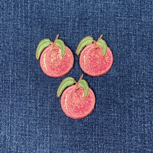Könnte beinhalten: Drei gestickte Pfirsich-Patches mit rosa Glitzer und grünen Blättern auf blauem Denim-Hintergrund. Die kreisförmigen Patches haben ein strukturiertes Aussehen, das auf ein Stoff- oder Filzmaterial hindeutet.
