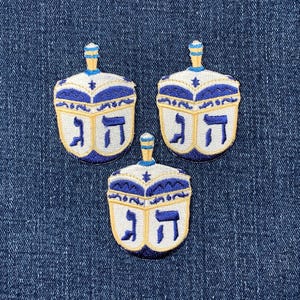 Könnte beinhalten: Drei blaue und weiße gestickte Bügeleisen-Aufnäher von Dreidels, einem traditionellen Kreisel, der während Chanukka gespielt wird. Jeder Dreidel hat die hebräischen Buchstaben 'Nun', 'Gimmel', 'Hey' und 'Shin' auf seinen Seiten.