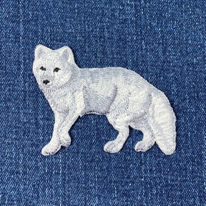 May include: An embroidered patch of an arctic fox, in white. The fox is standing on all four paws with its tail curled behind it.