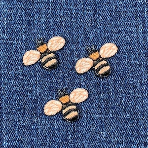 May include: Three embroidered bee patches on a blue denim background. The bees are yellow and black with brown wings.