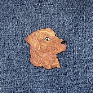 May include: Embroidered patch depicting a golden retriever dog's head in profile. The patch uses shades of brown and tan, with detailed stitching. It is set against a blue denim background, creating a contrast in textures and colours.