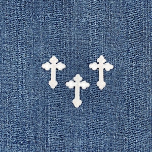 Könnte beinhalten: Drei weiße, kreuzförmige Applikationen mit dekorativem Rand sind auf einem blauen Jeansstoff-Hintergrund angeordnet. Die Kreuze sind gleichmäßig verteilt und scheinen aus einem strukturierten Material zu bestehen.