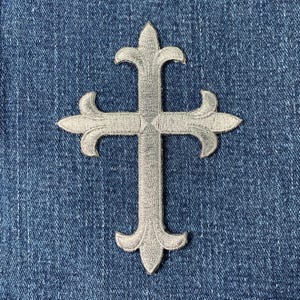 May include: A silver-toned embroidered cross patch featuring fleur-de-lis style ends on each of the four arms. The cross has a detailed stitched texture and is centred against a dark blue denim fabric background with a visible twill weave.