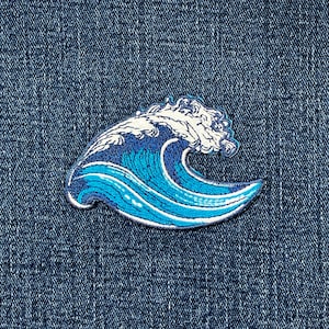 May include: A blue and white embroidered patch featuring a stylized wave design. The wave is depicted with shades of blue and white, with a detailed crest and flowing lines. The patch is likely for clothing or accessories.
