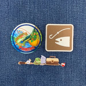 May include: Three embroidered patches featuring fishing themes. One circular patch shows a person in a yellow boat catching a fish with the word FISHING. One square brown patch depicts a white fish and hook. One long patch shows gear.