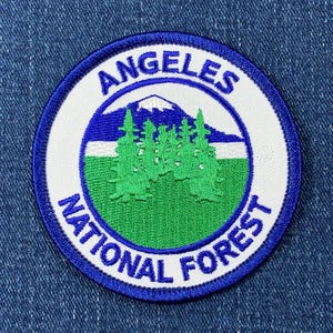 May include: A white and blue embroidered patch with the text "Los Angeles National Forest" and a green and blue illustration of a mountain range with trees.