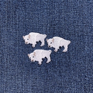 May include: Three white bison patches are arranged on a blue denim background. Each patch is embroidered with detailed features, including the bison's body, legs, and head. The patches are small and suitable for clothing or craft projects.