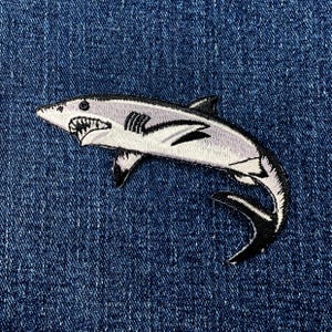 May include: Embroidered shark patch featuring a gray and white shark with black accents, including the fins and tail. The shark has a detailed design with visible teeth and is set against a blue denim background.