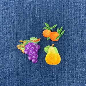 May include: Three embroidered fruit patches featuring a cluster of purple grapes with green leaves, a bright orange citrus fruit with green leaves, and a yellow pear with a green leaf, all displayed against a dark blue denim fabric background.
