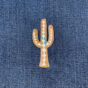 May include: A tan and white cactus patch with a turquoise and orange design. The patch is embroidered and has a detailed, textured appearance. The cactus is set against a blue denim background.