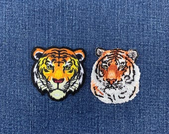 May include: Two embroidered tiger head patches. The left patch features a vibrant orange, yellow, and black tiger face with a thick black border. The right patch displays a detailed tiger head in shades of orange, white, and black.