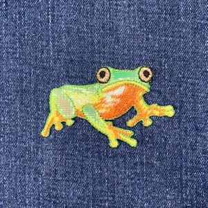 May include: Green and orange embroidered frog patch on a blue denim background.