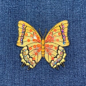 以下が含まれることがあります： 青いデニム生地に刺繍された、黄色とオレンジの虹色のスパンコールが付いた蝶々です。