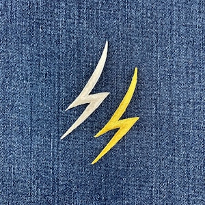 May include: Two embroidered lightning bolt patches, one in a metallic silver thread and one in a vibrant yellow thread, are positioned side by side against a dark blue denim fabric background with a visible woven texture.