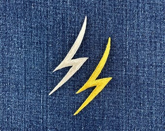 May include: Two embroidered lightning bolt patches, one in a metallic silver thread and one in a vibrant yellow thread, are positioned side by side against a dark blue denim fabric background with a visible woven texture.
