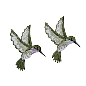 May include: Two embroidered hummingbird patches featuring olive green wings and tails with white feather detailing. Each bird has a slender black beak and a small patch of light purple colour on the throat area, captured in a flying pose.