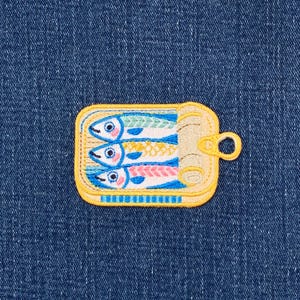 May include: Embroidered patch of a yellow sardine tin, containing three colourful fish. The fish have blue, green, pink, and yellow details. The patch is set against a blue denim background.