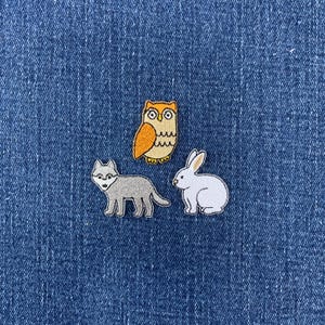 May include: Three embroidered patches featuring a tan and orange owl, a gray wolf standing in profile, and a white rabbit sitting. Each patch has a detailed stitched border and is displayed against a textured blue denim fabric background.