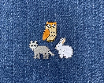 May include: Three embroidered patches featuring a tan and orange owl, a gray wolf standing in profile, and a white rabbit sitting. Each patch has a detailed stitched border and is displayed against a textured blue denim fabric background.