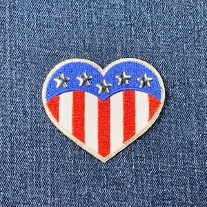 May include: A red, white, and blue heart-shaped patch with stars and stripes. The patch is made of fabric and has a white outline.