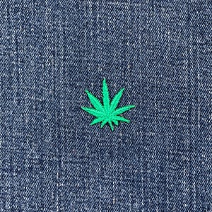 Könnte beinhalten: Ein kleiner, hellgrüner, gestickter Cannabisblatt-Aufnäher auf einem dunkelblauen Jeanshintergrund. Der Aufnäher ist zentriert und detailliert, mit sichtbaren Nähten. Der Denim hat ein strukturiertes Aussehen.