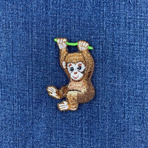 May include: An embroidered patch featuring a brown monkey with white facial details and hands, hanging from a bright green branch. The monkey has a light brown chest and darker brown fur on its back and limbs, rendered in detailed thread work.