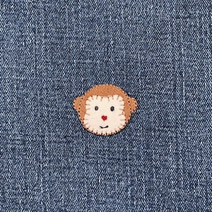 May include: A felt monkey face applique with a brown head and ears, a cream-colored face, and a red heart-shaped nose. The applique is stitched onto a blue denim background.