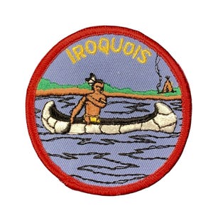 May include: A circular embroidered patch with a red border featuring a light blue background. The design depicts a person in a white canoe on blue water, with green land, a small teepee, and the yellow text IROQUOIS arched at the top.