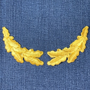 May include: Two gold embroidered leaf patches, each with detailed veining, are arranged in a curved shape against a blue denim background. The patches are likely for decorative purposes, such as embellishing clothing or accessories.