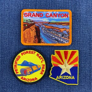 May include: Three embroidered patches featuring a rectangular Grand Canyon landscape, a circular Petrified Forest National Park patch with a log graphic, and a shield-shaped Arizona state flag patch with a gold star and the text ARIZONA.