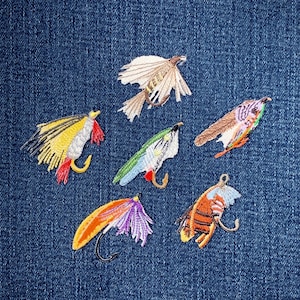 Könnte beinhalten: Gestickte Angelköder in verschiedenen Farben, darunter Gelb, Rot, Grün und Orange, mit detaillierten Nähten. Jeder Köder hat einen gebogenen Haken und befindet sich auf einem blauen Jeanshintergrund.