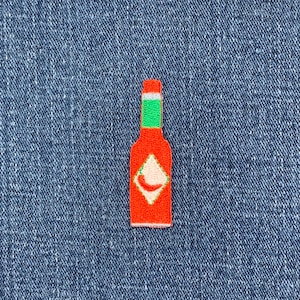 May include: Embroidered patch of a red hot sauce bottle with a green neck and white diamond-shaped label featuring a red chili pepper. The patch is on a blue denim background.