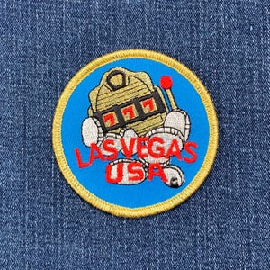 May include: A blue and gold embroidered patch with a cartoon slot machine and the text "Las Vegas USA".