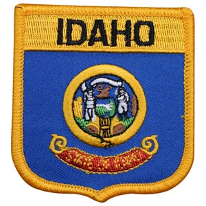 May include: An embroidered patch featuring the state seal of Idaho, with the words "IDAHO" in black lettering, on a blue background with a gold border.