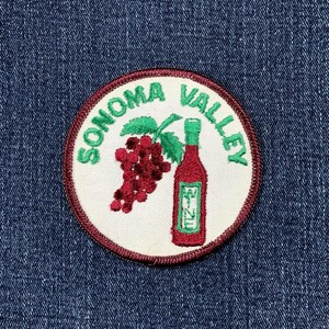 May include: Embroidered patch with a white background, featuring the words "SONOMA VALLEY" in green, surrounding a red wine bottle and a bunch of grapes. The patch has a burgundy border and is set against a blue denim background.