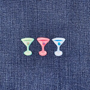 May include: Three embroidered cocktail glass patches in green, pink, and blue, each with a small dot in the center. The patches are set against a textured, dark blue denim background.