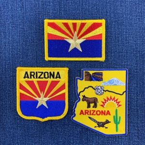 May include: Three embroidered patches featuring Arizona themes. One rectangular patch shows the Arizona state flag. One shield-shaped patch displays the Arizona flag with the text ARIZONA. One map-shaped patch depicts a desert scene with a cactus, a donkey, a roadrunner, a mountain, a canyon, and a Route 66 sign.