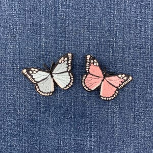 May include: Two embroidered butterfly patches featuring detailed black stitching along the wings. One butterfly is light blue with white accents, and the other is soft pink with white accents. Both patches have black borders and antennae.