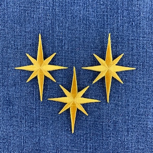 May include: Three embroidered patches in the shape of eight-pointed stars. Each star is a vibrant golden yellow colour with a dense, stitched texture. The patches are arranged in a triangular formation, featuring sharp, elongated points.