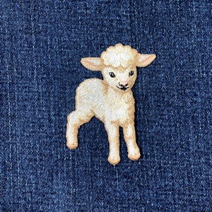 May include: Embroidered patch of a cream-coloured lamb with a fluffy head, standing on four legs. The lamb has black eyes and a black nose. The patch is set against a dark blue denim background.
